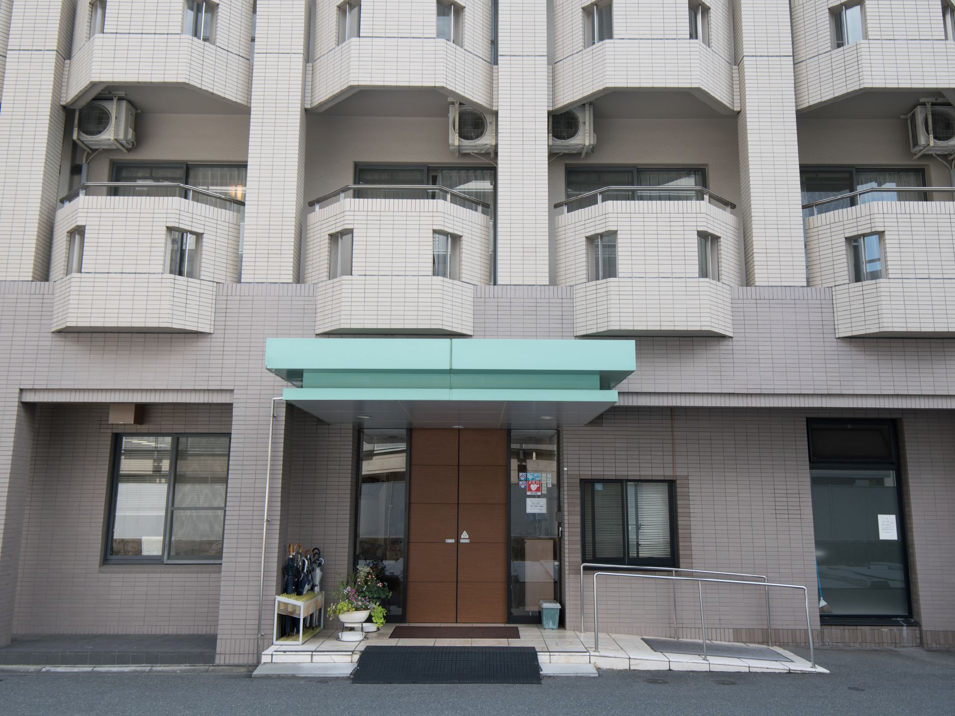施設の出入り口は茶色の自動ドアになっている。ちょっとした段差がありスロープや手すりが設置されている。傘立てや花も置かれている。