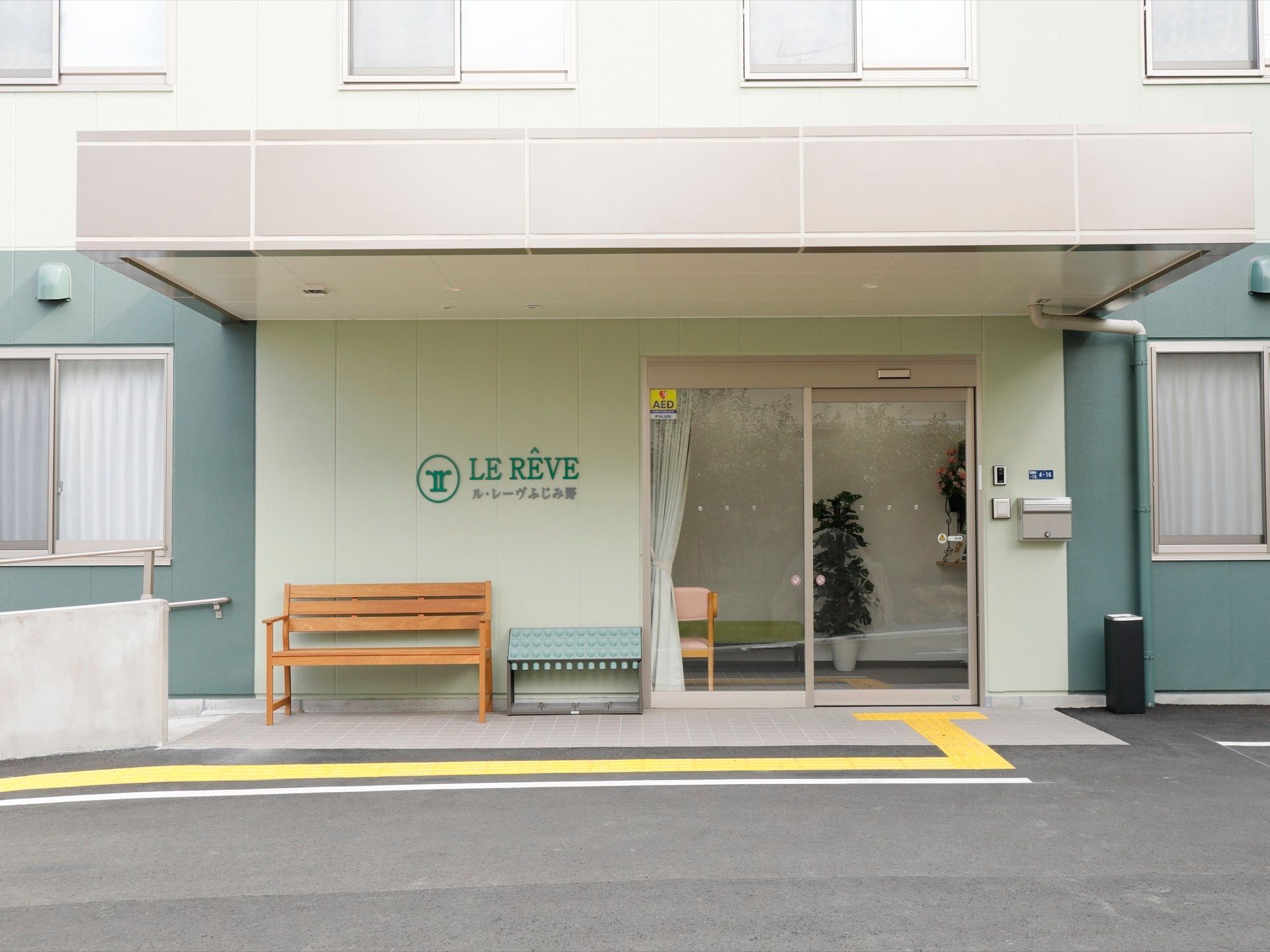 薄いグリーンの爽やかな外観の施設で、玄関は自動ドアになっている。ドアの横にはインターホンとポストが設置されている。木製ベンチが置かれている。