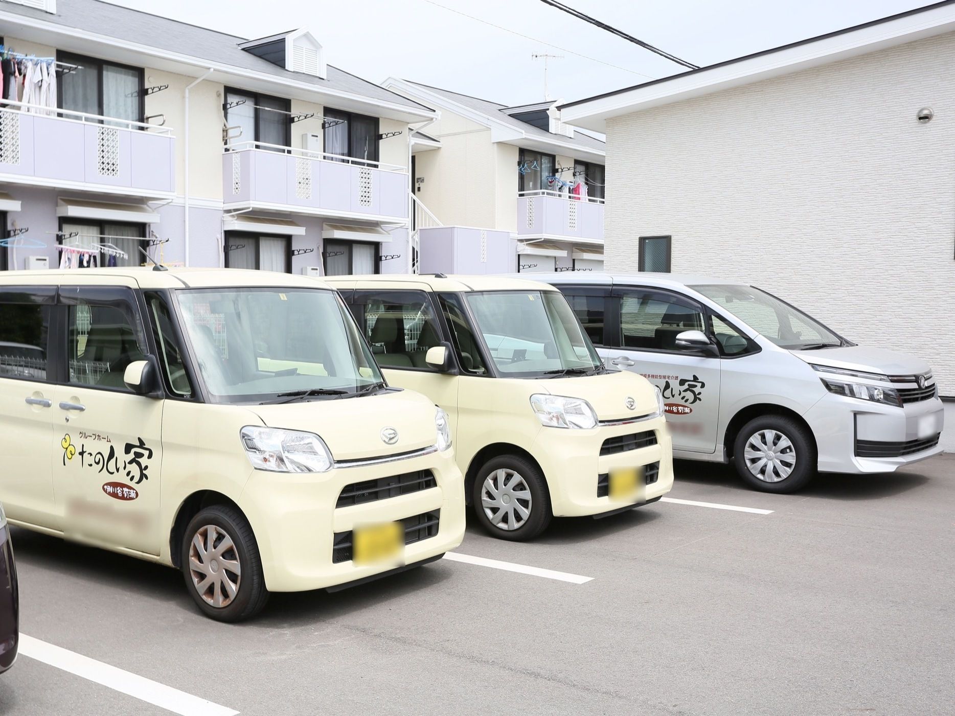施設が完備する駐車場には、施設の送迎車が３台停車している。クリーム色の車が手前に２台、次いでシルバー色の車が停まっている。