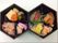 ６角形の２段弁当箱に盛り付けられた食事である。赤飯に煮物、かまぼこ、刺身などおかずが７種。黒の器によく映えて食欲をそそる。
