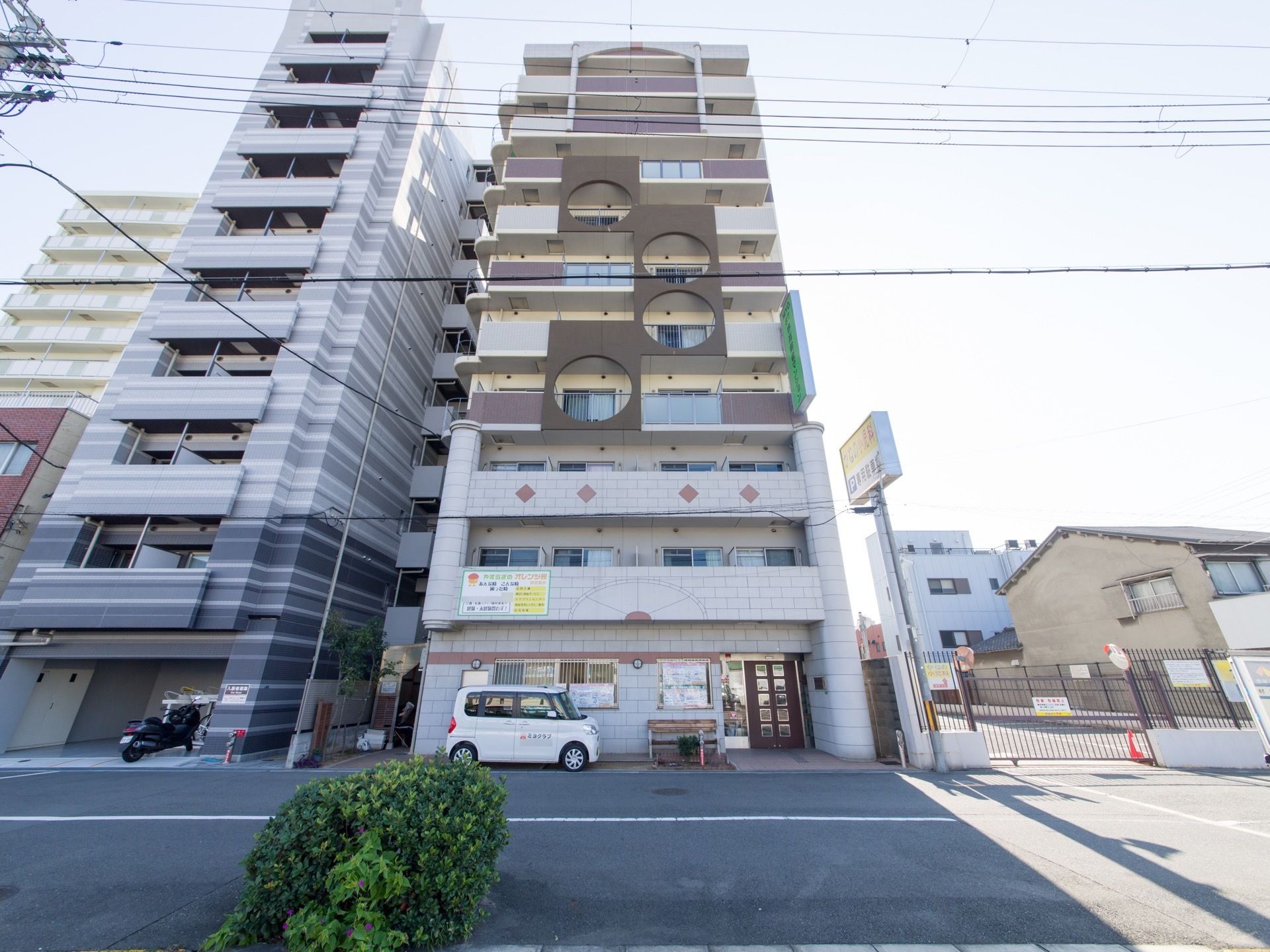マンションタイプの施設である。隣地との境の樹を植えてある。施設用の車を建物に沿って駐車してある。外のための照明が外壁に付けられている。