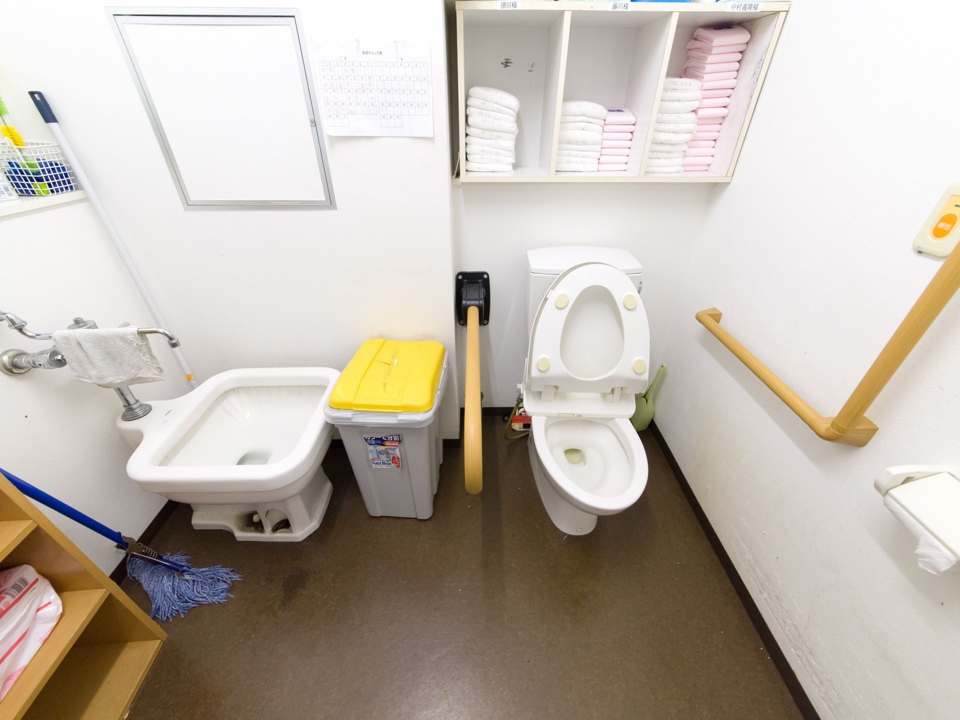 トイレの壁に向かって右側に温水洗浄便座、その左に大型のごみ箱、大型の汚物流しがある。壁の棚に入居者様毎に備品が置かれている。