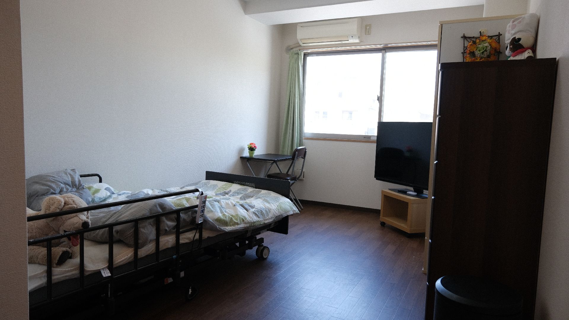 寝具付きのベッドが置かれたフローリングの部屋には大きな窓があり、テレビや家具も置かれている。