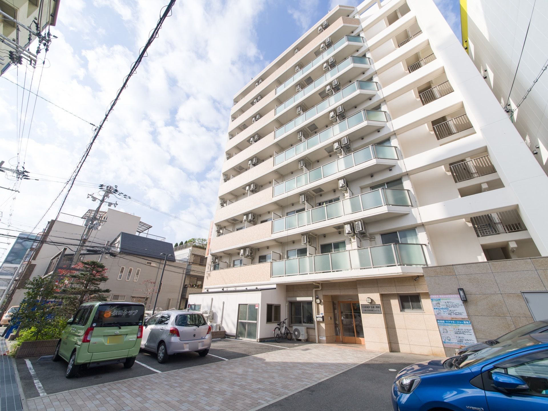 施設は9階建てである。玄関入口の両側はコンクリートの平地で、車の駐車スペースとして利用されている。4台の車が止められている。