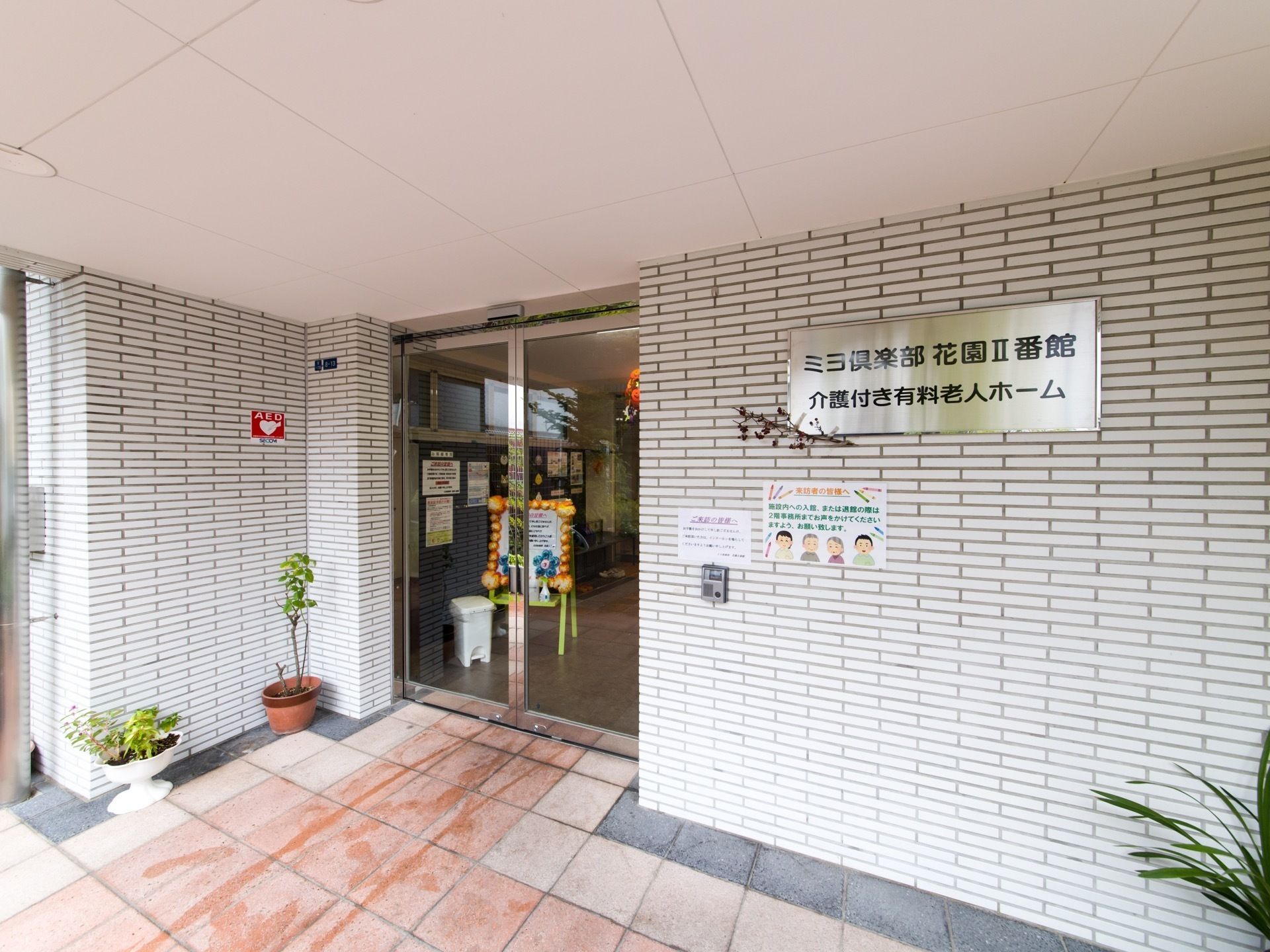 入口前の壁には、AEDを設置してあることを示すシールがある。ガラスドアの向こうには、来訪者様へのメッセージボードなどが内部が見えている。
