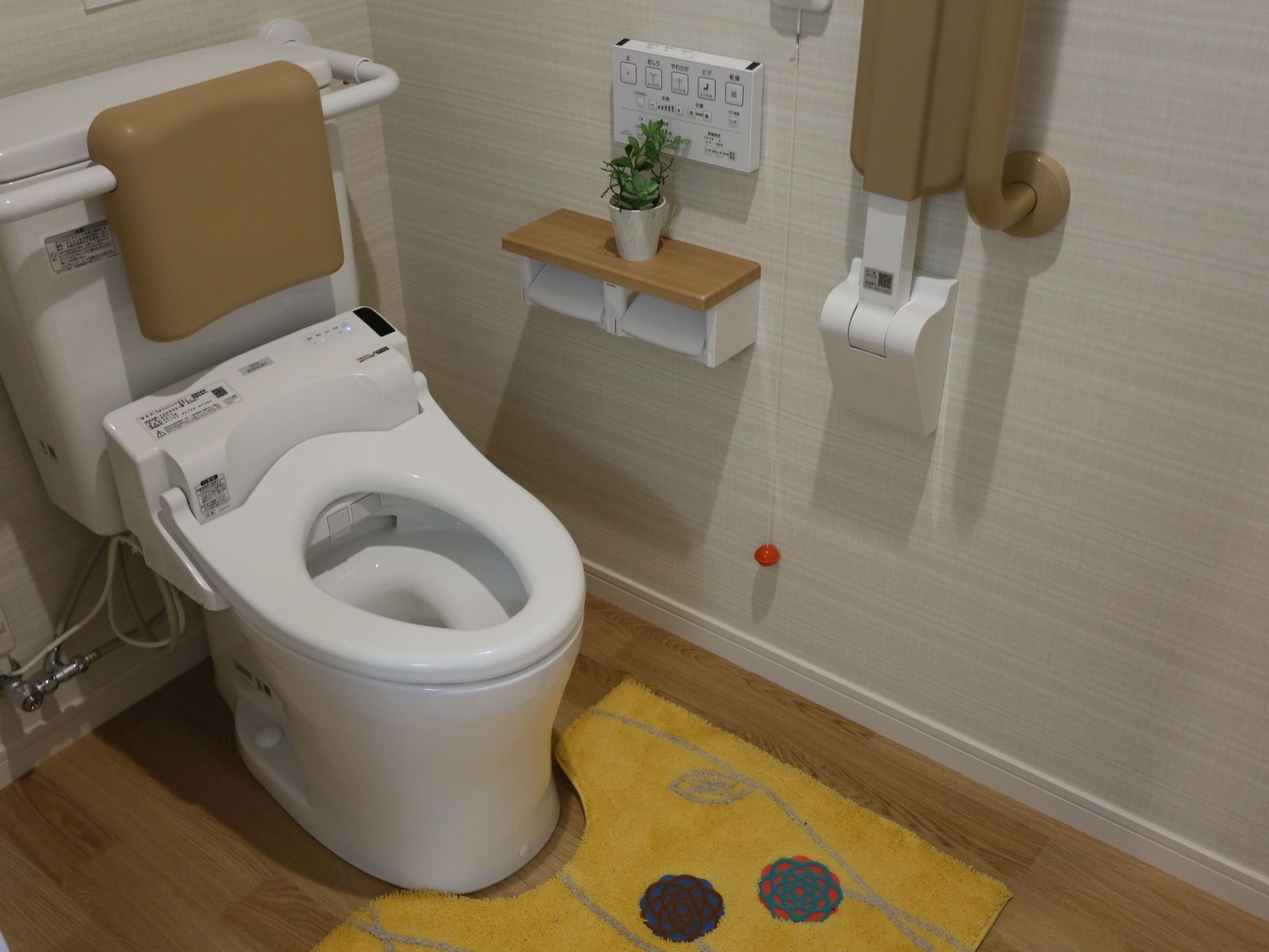 温水洗浄便座付きトイレである。背面のタンクに背もたれクッションがある。左手に操作スイッチ、ペーパーホルダーと緊急ボタンがある。