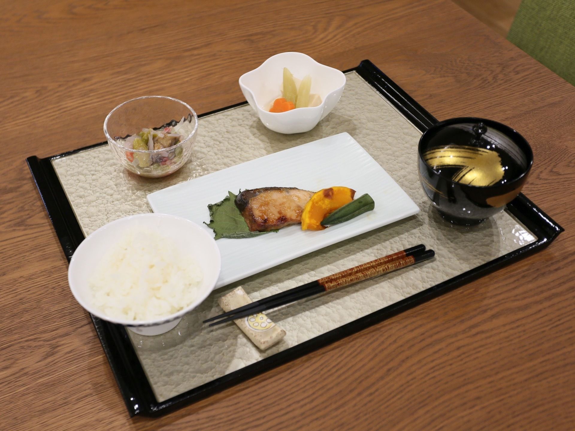 お盆の上にご飯、汁物、焼き魚、小鉢、お箸などが置かれている。お椀やお箸など高級感がある。木目調のテーブルの上に配膳されている。