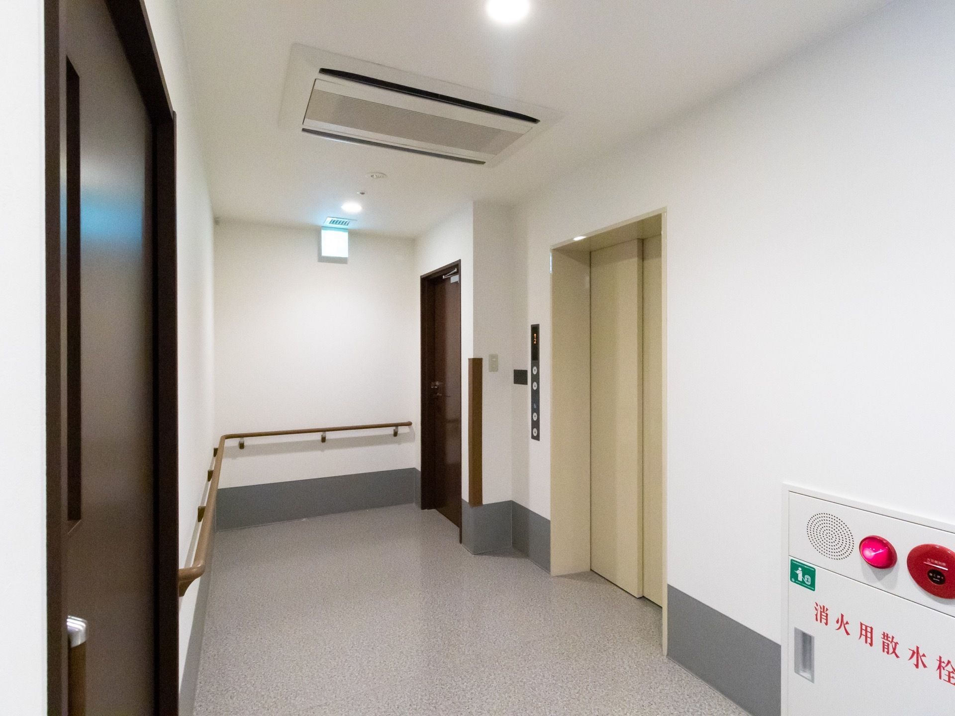 白い壁とグレーの床の廊下がある。居室のドアとエレベーターがある。壁には手すりが設置されており、消火栓がある。