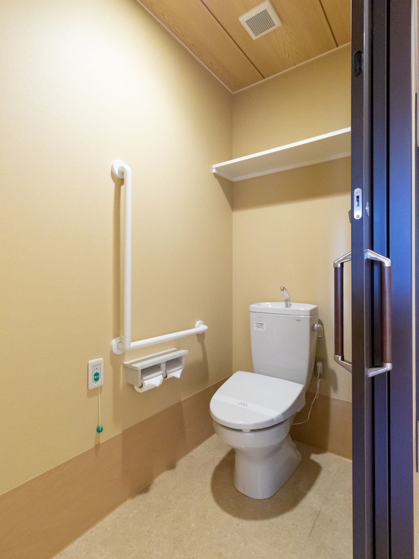 クリーム色の壁に囲まれた個室のトイレがある。引き戸のドアがあり、壁には手すりが設置されている。天井付近には棚がある。