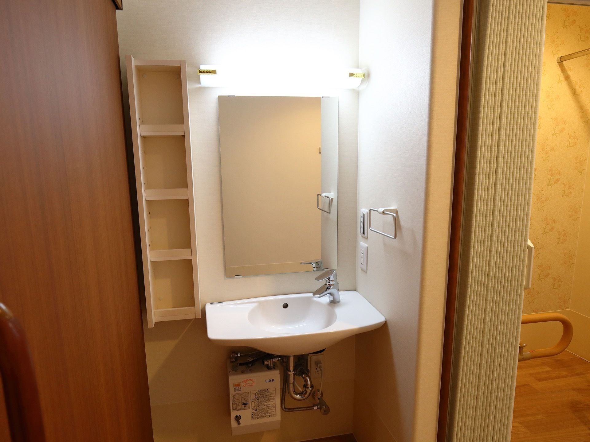 白色の壁に鏡を設け、横に４段に分かれた細長い棚を取り付けている。下に洗面ボウルを設置し、横側にタオル掛けを備えている。