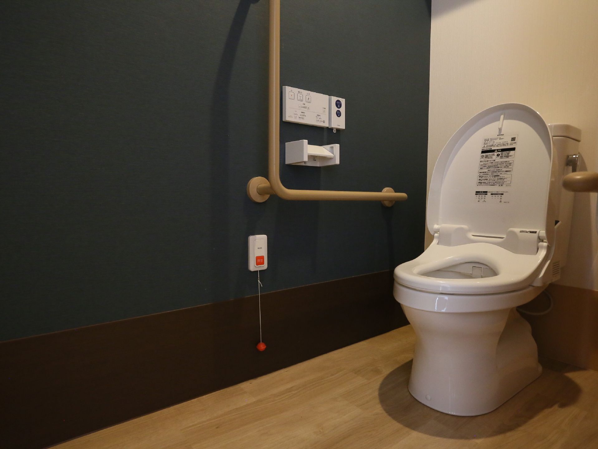 ダークグリーンの壁が印象的なトイレである。温水洗浄便座やＬ型手すり、呼出ボタンが完備されている。壁には、便器洗浄用リモコンも設置されている。