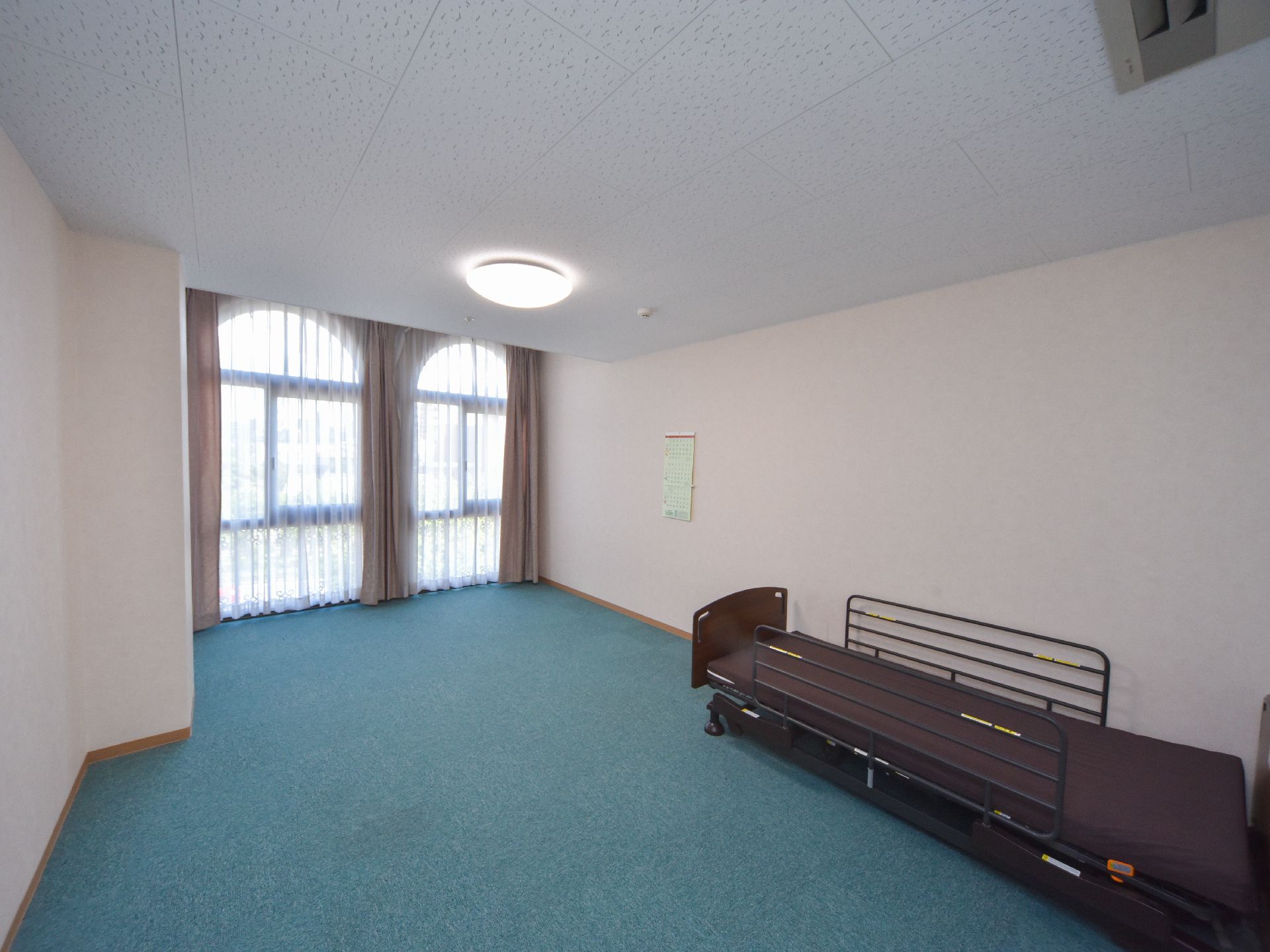 居室は快適に過ごせる十分な広さが確保されており、床には青系の絨毯が敷かれている。壁際に介護ベッドが置かれている。