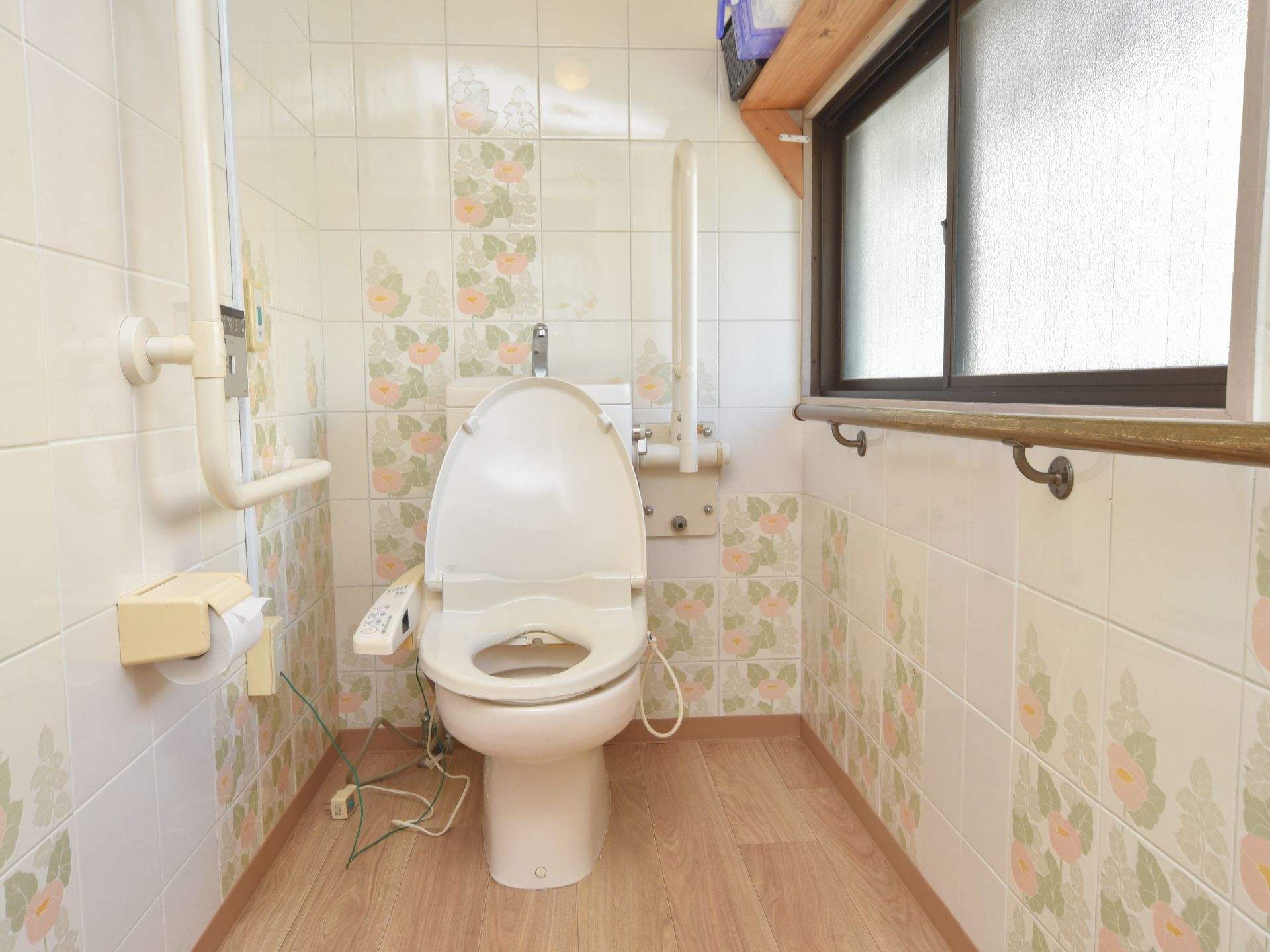 トイレの壁には花柄のタイルが使われている。L字型や可動式の手すり、緊急呼び出しボタンが設置されている。