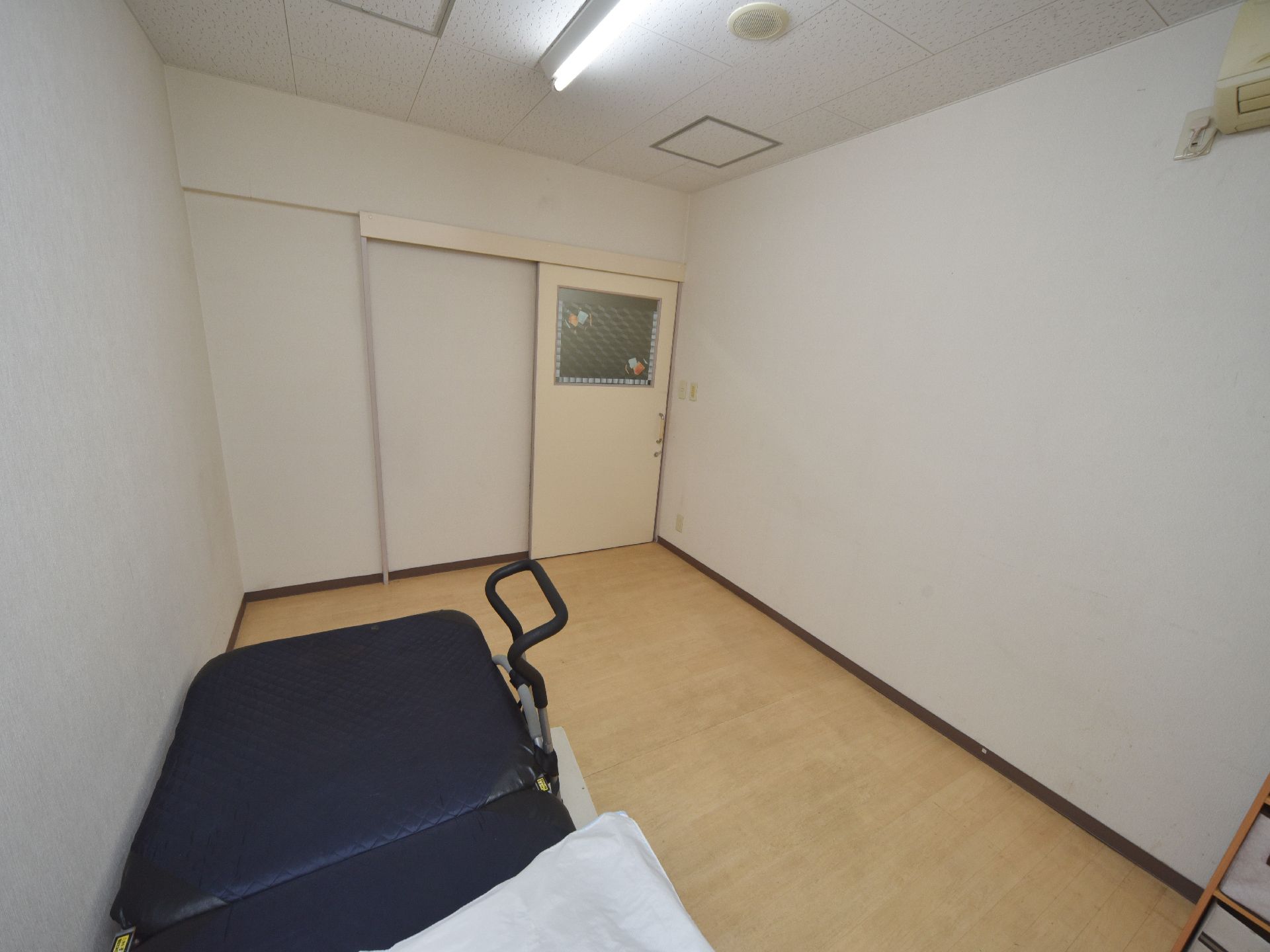 居室のドアは引き戸になっている。壁際にベッドが置かれおり、立ち上がり用のアームが取り付けてある。エアコンが設置されている。