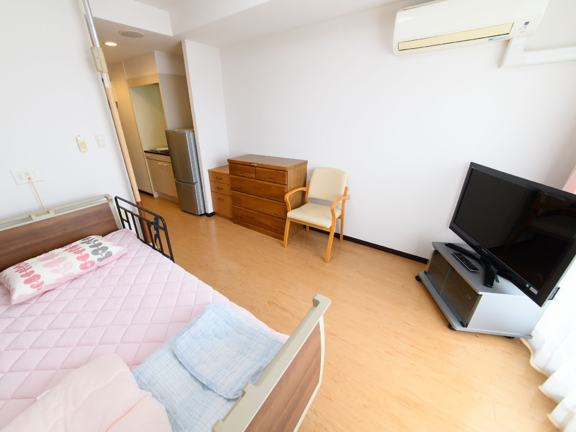 ワンルームのフローリングの部屋で、廊下部分には冷蔵庫、キッチンが見える。電動ベッドには柵があり、布団などが落ちないようになっている。