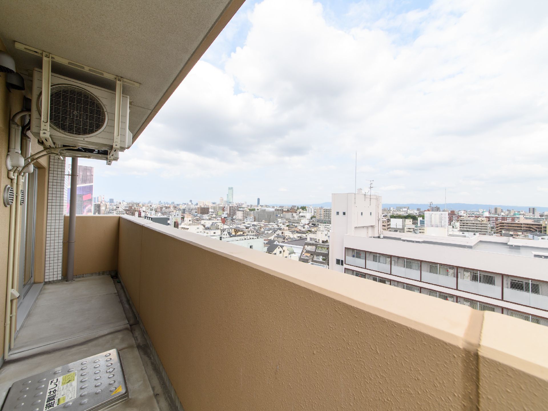 茶色の塀で囲まれたベランダがあり、室外機が置いてある。目の前には高層マンションがあり、屋上にアンテナが設置してある。