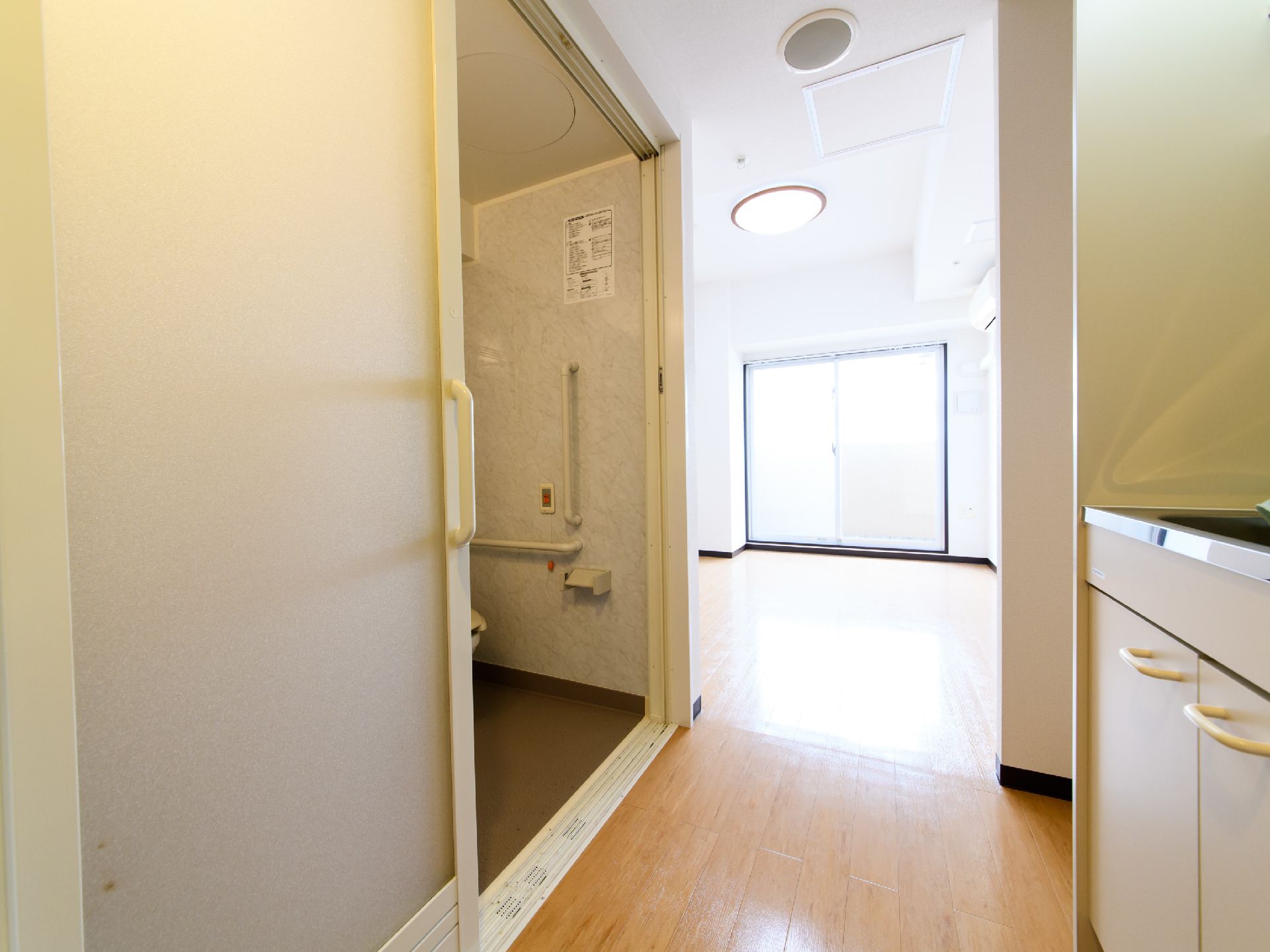 引き戸が半分開いていて、洋式トイレが見える。壁には手すりと緊急時呼び出しブザーが設置してある。ワンルームで、掃き出し窓がついている。