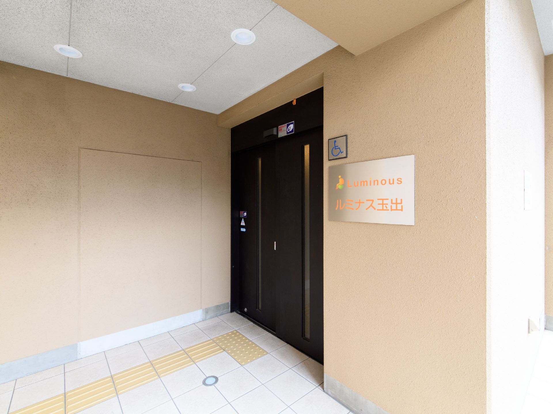 天井に埋め込み式の照明があるが､点灯はしていない玄関前で、壁に施設の名前が書いてある看板が取り付けてある。