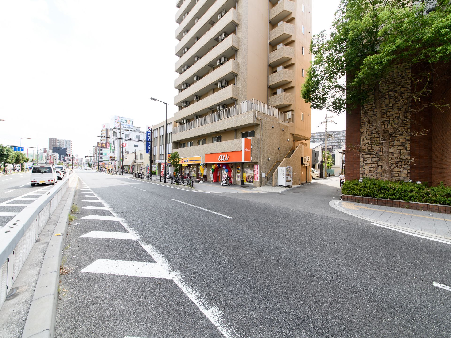 施設前の道路は、片道二車線と三車線に分かれた大きな道路があり、車が6台ほど見える。施設の階下はテナントとなっており商業施設が入っている。