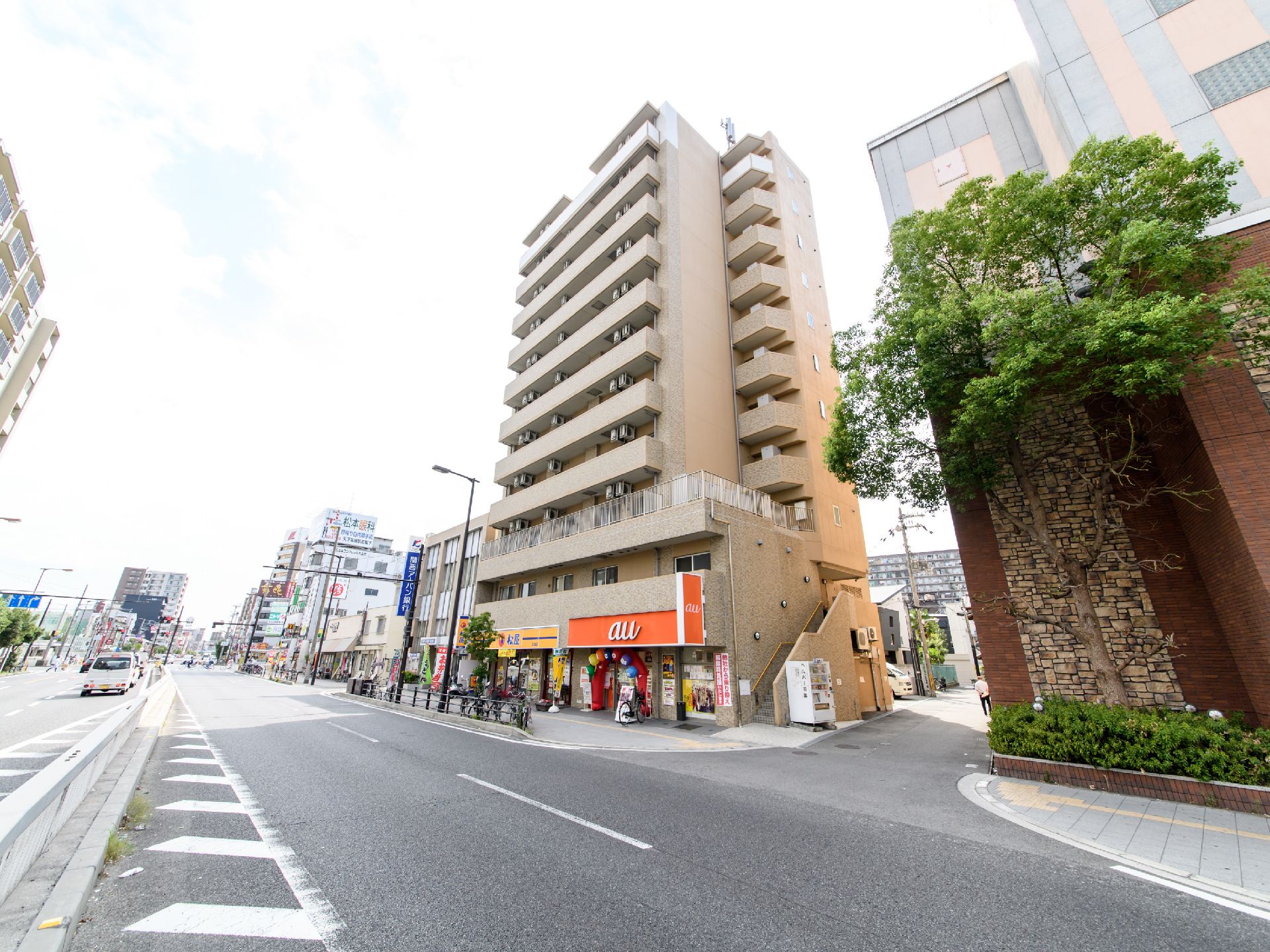 二車線と三車線の大きな道路が見える。車が数台赤信号のため停車しており歩行者が見える。施設の外階段そばに自動販売機が置いてある。