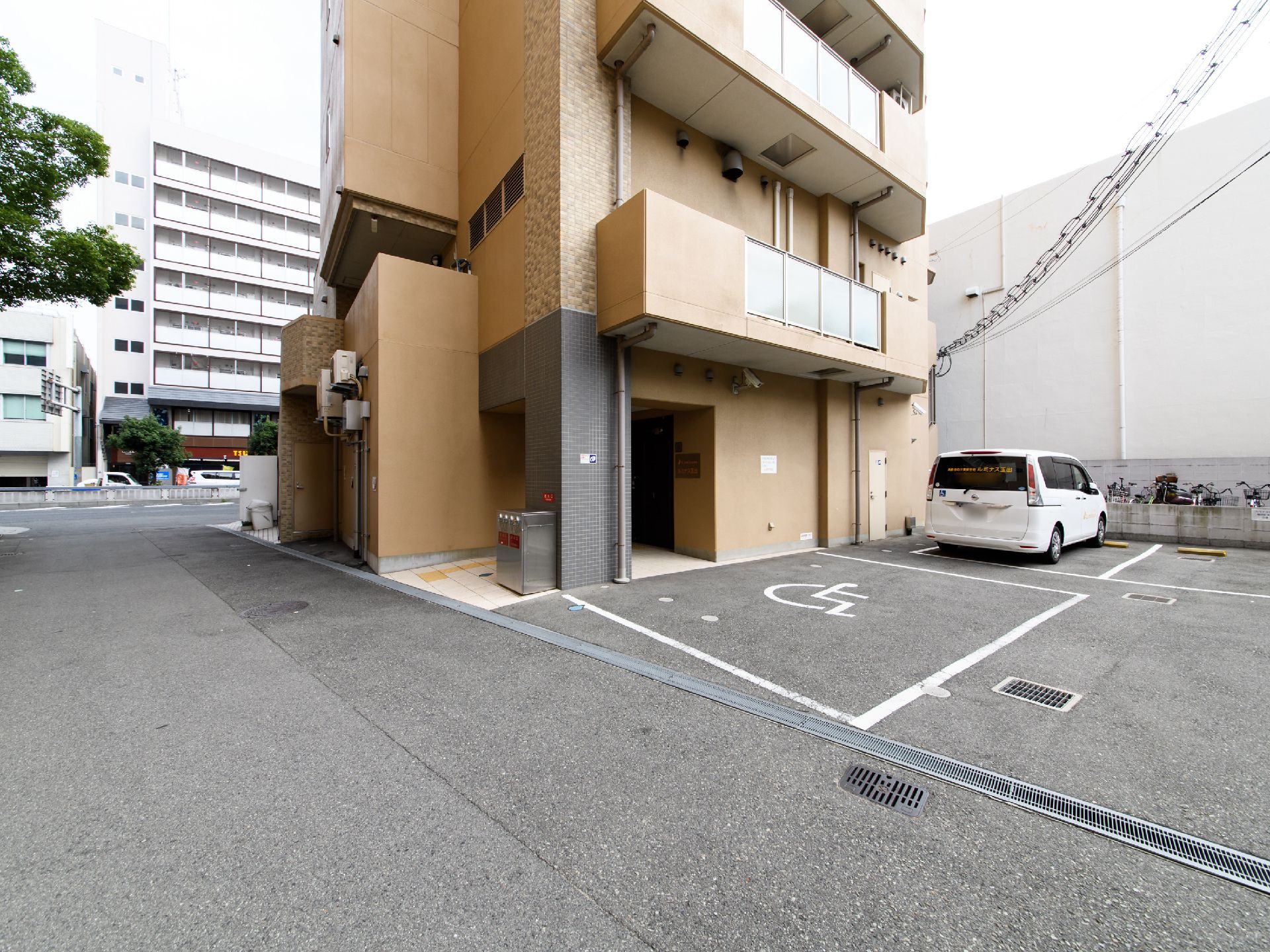 施設の敷地内には、駐車場が設けてあり、身障者マークがついている個所もある。施設の建物は全体的に茶色く、ベランダに塀があるのが見える。