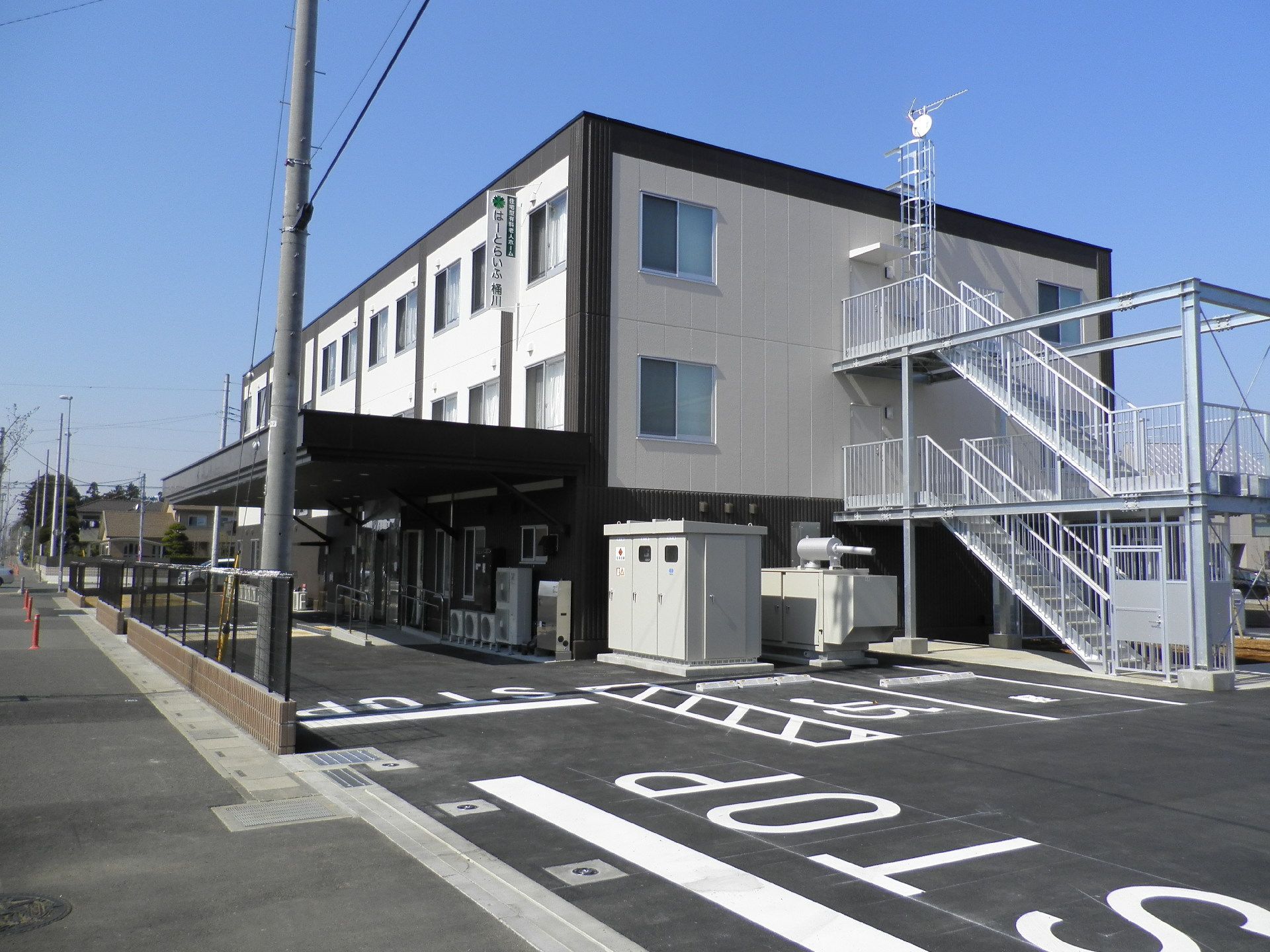 施設の建物の横には駐車場と、大きな外階段が設置されている。クリーム色と茶色の外観で、すっきりとした印象を与える建物になっている。