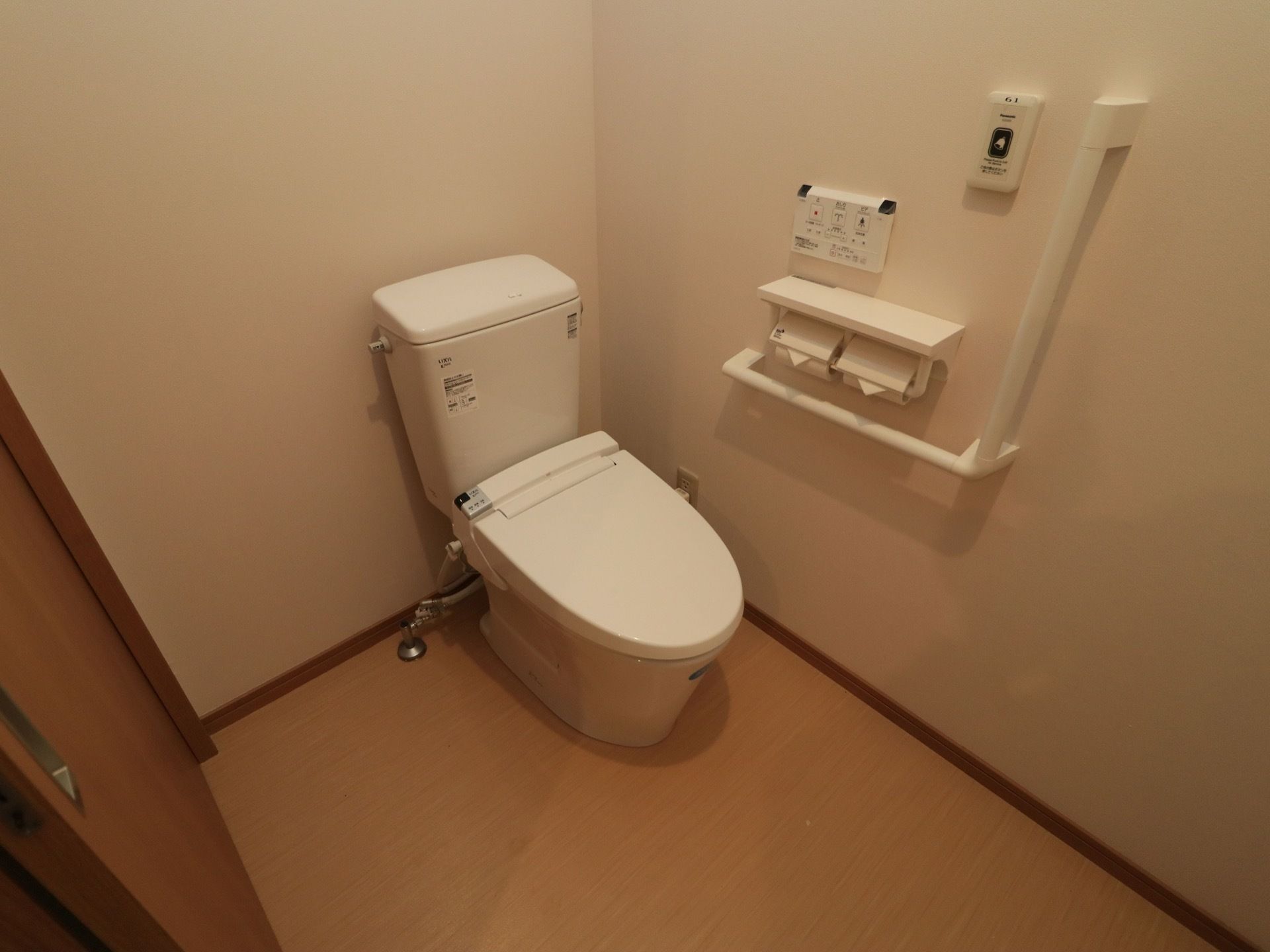 フローリングと木製のドアの個室のトイレ。壁には縦横方面に手すりが設置されており、温水洗浄便座がついている。