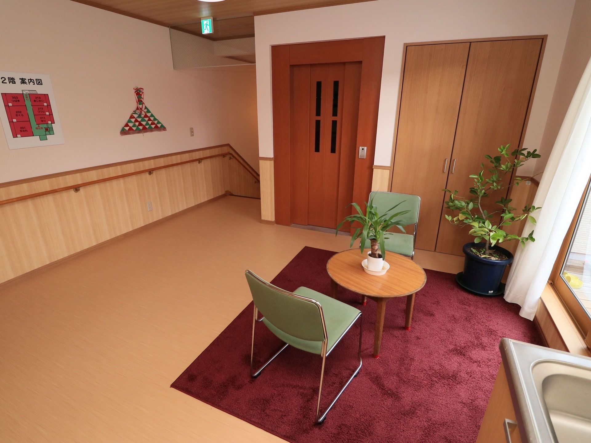 茶色のドアのエレベーターが設置してあるホール。ワインレッドのラグマットが敷かれた一角にはテーブルと椅子が置かれている。