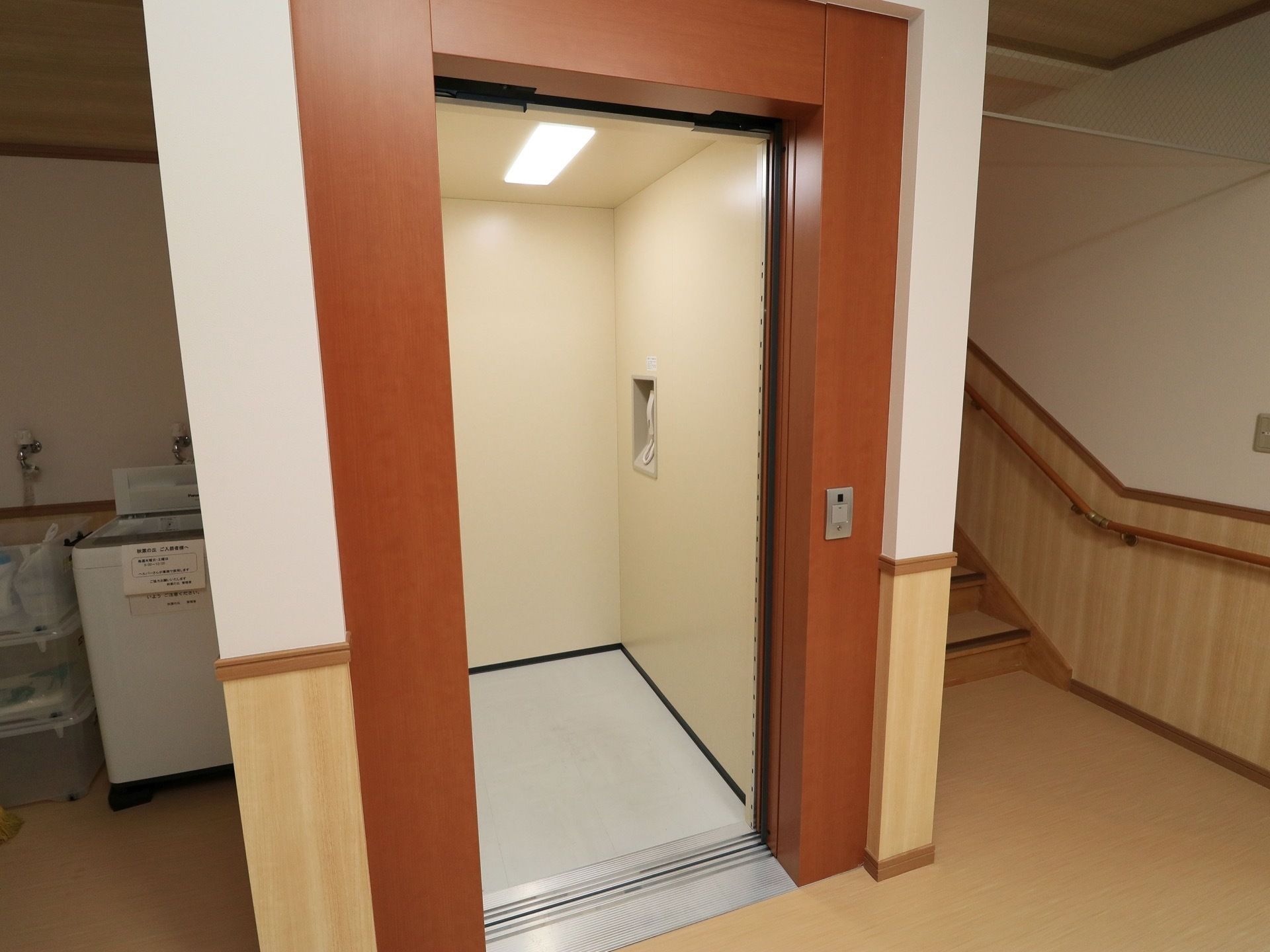 茶色い枠のエレベーターが1基設置されている。中の壁側には電話の受話器がある。上には蛍光灯がつけられている。