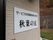 クリームがかった色の外壁がある。そこには白い看板が設置されている。看板には黒色で施設名が書かれている。