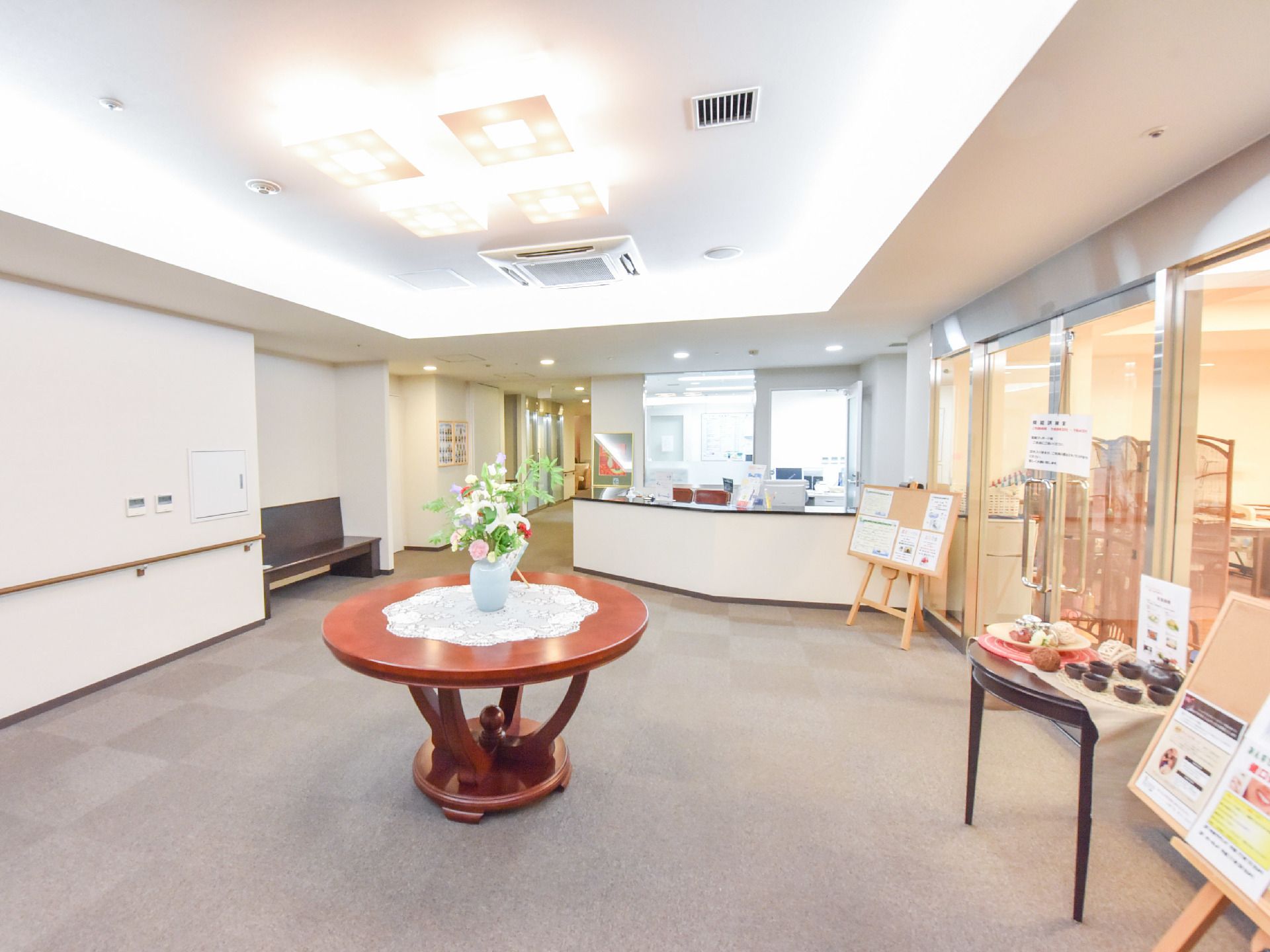 ガラス張りの部屋の前にホールがある。天井にはおしゃれでゴージャスな照明がある。ベンチがあり、作品や花が飾られている。