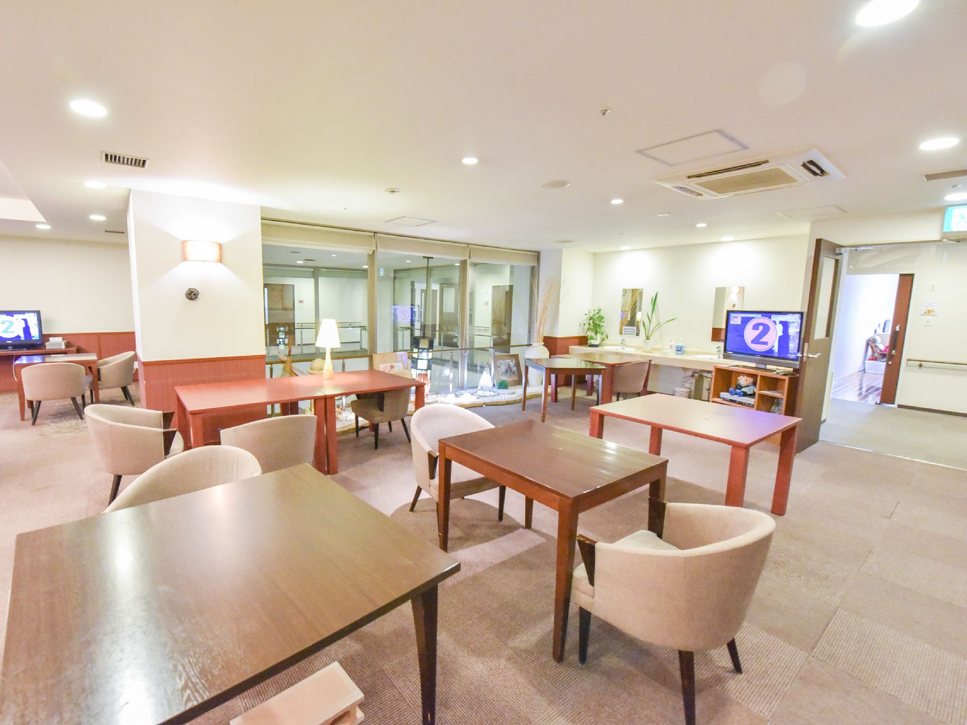 "明るくて広い部屋に2人ずつ座れるようテーブルセットが配置されている。テレビが2台とエアコンがある。"