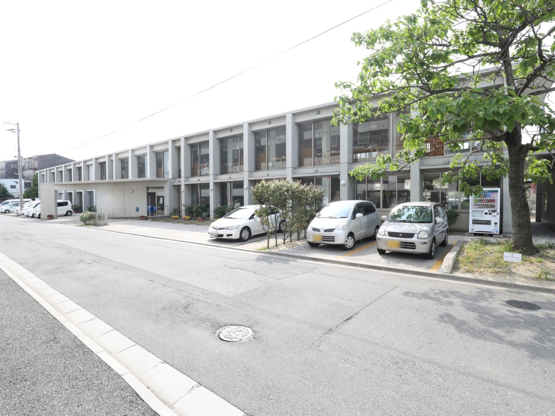 施設正面には大きな木が１本植えてあり、その隣に駐車場が設けてある。並列式の駐車場で、植え込みで車と車の間を間仕切りしてある部分がある。