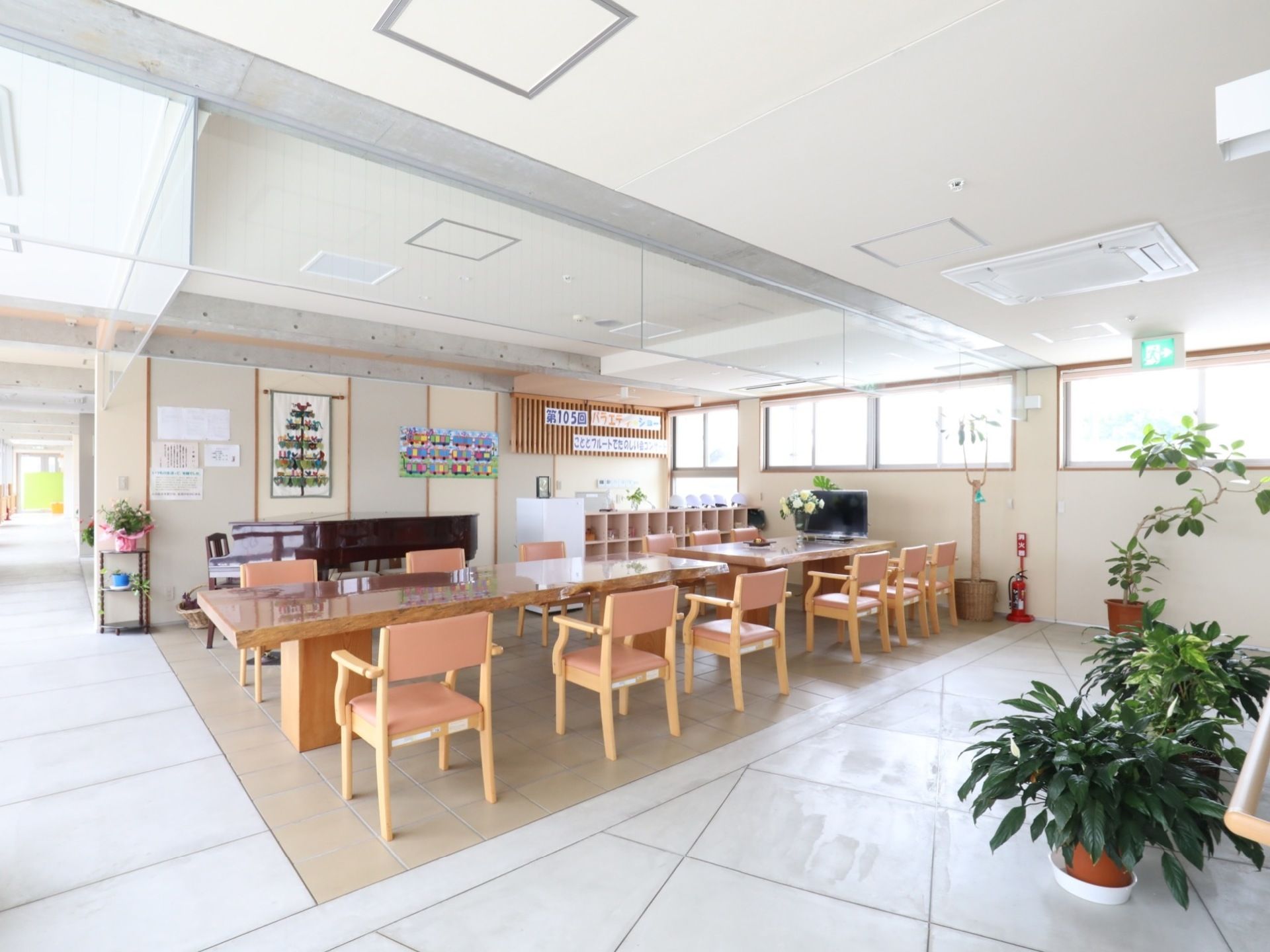 広々としたスペースに椅子とテーブルが複数設置してある。手前には観葉植物が置いてあり、奥の壁手前にはグランドピアノが置いてある。