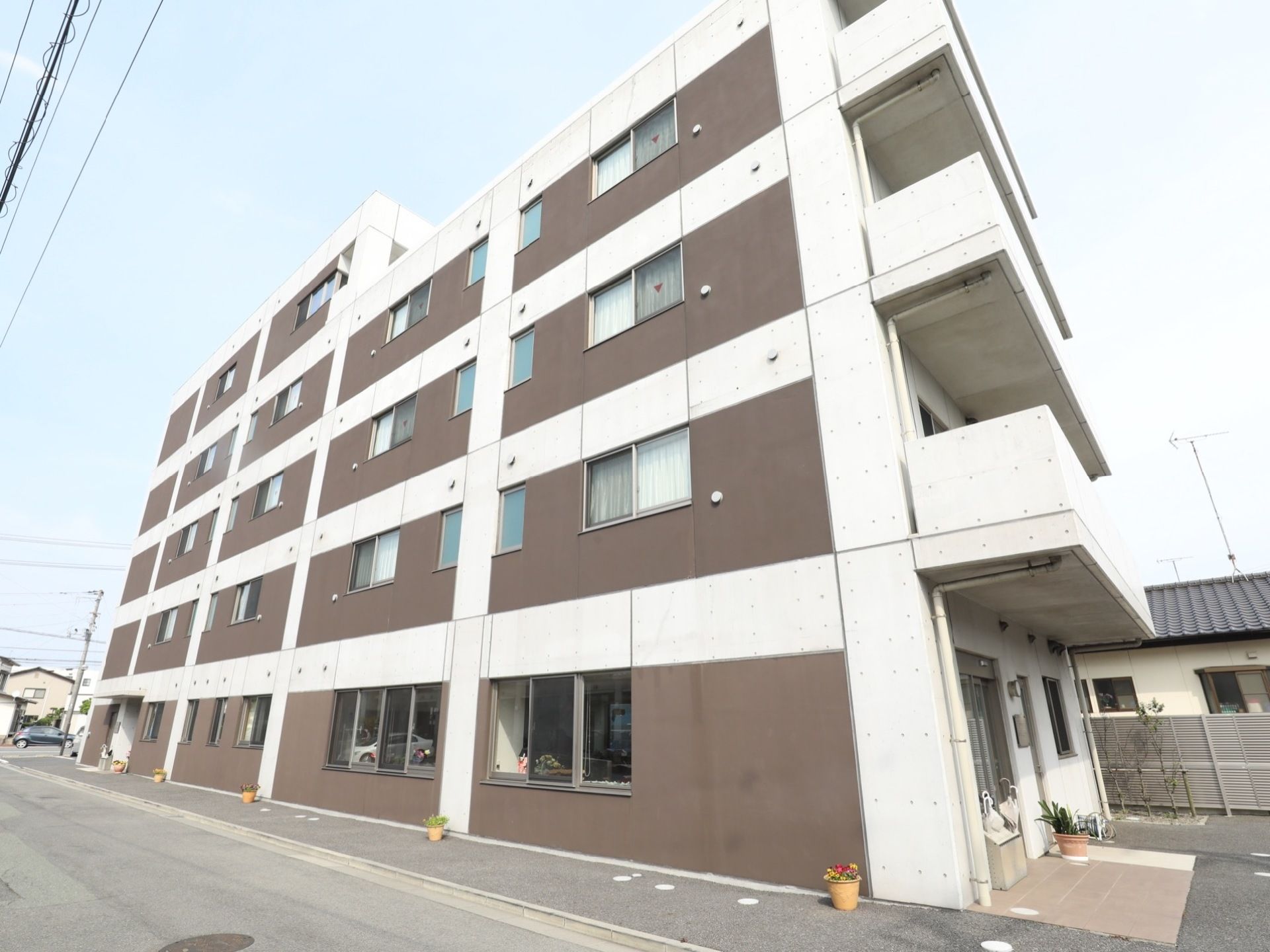 壁は茶色と白色のシックな雰囲気で、５階建ての建物である。建物の周りや玄関には、鉢植えの花が飾られている。