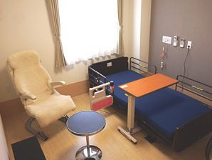 居室には、窓の前に専用テーブルが付いた介護用ベッドがある。寛ぎスペースには、背もたれ付きのリクライニングチェアと丸テーブルが置いてある。