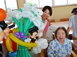 椅子に座った高齢女性とカラフルなバルーンアートを持ったスタッフ