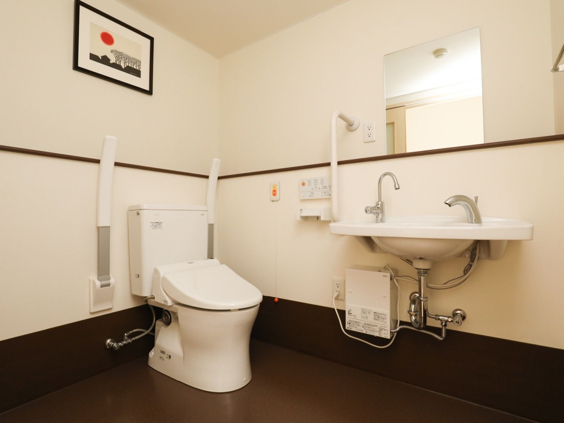 ダークブラウンの床のトイレスペースである。便座の両側に可動式の手すりを備え、横側に洗面台を設置している。天井には絵を飾っている。