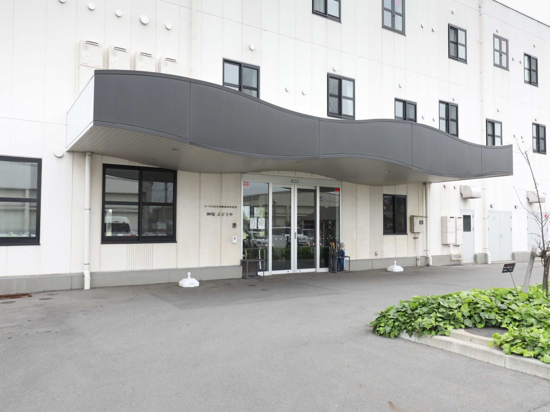 ガラス張りの玄関ドアの前方に大きなカーブを描いた屋根を設けている。正面には緑豊かな花壇を設けている。