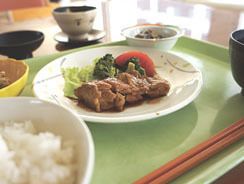 モスグリーンのトレーには、中央にメインディッシュの肉料理にトマトと野菜が添えられたお皿がある。脇には小鉢やご飯、味噌汁などが並んでいる。