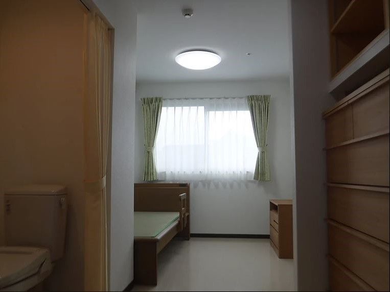 居室はゆったりと広さがあり、部屋の奥には緑色のカーテンの掛けられた窓が1つ設置されている。天井にはシーリングライトが取りつけられている。