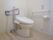 フロアの角部分にトイレが設置されている。便器の両側には白い手すりがあり、壁には引き紐付きのナースコールがある。