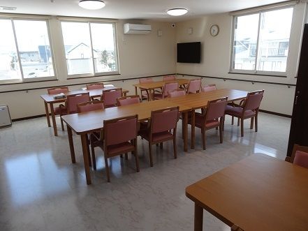 窓がある明るい空間に、テーブルとひじ掛け付きの椅子が並んでいる。天井と床が白色系で統一されていて、テレビやエアコンも設置されている。