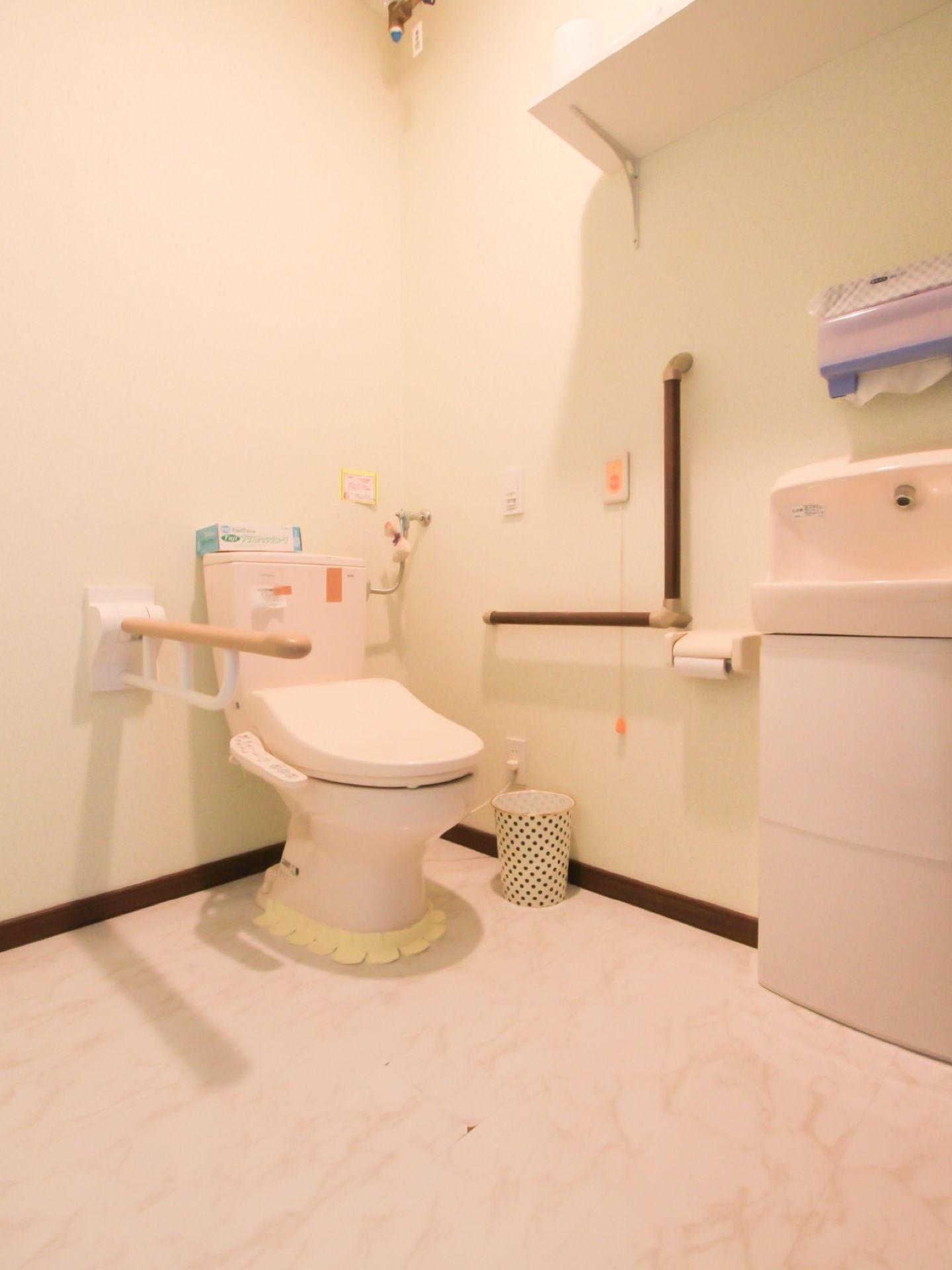 床と壁が白で統一された個室トイレである。中央に温水洗浄便座が取り付けられており、壁に手すりが設置されている。