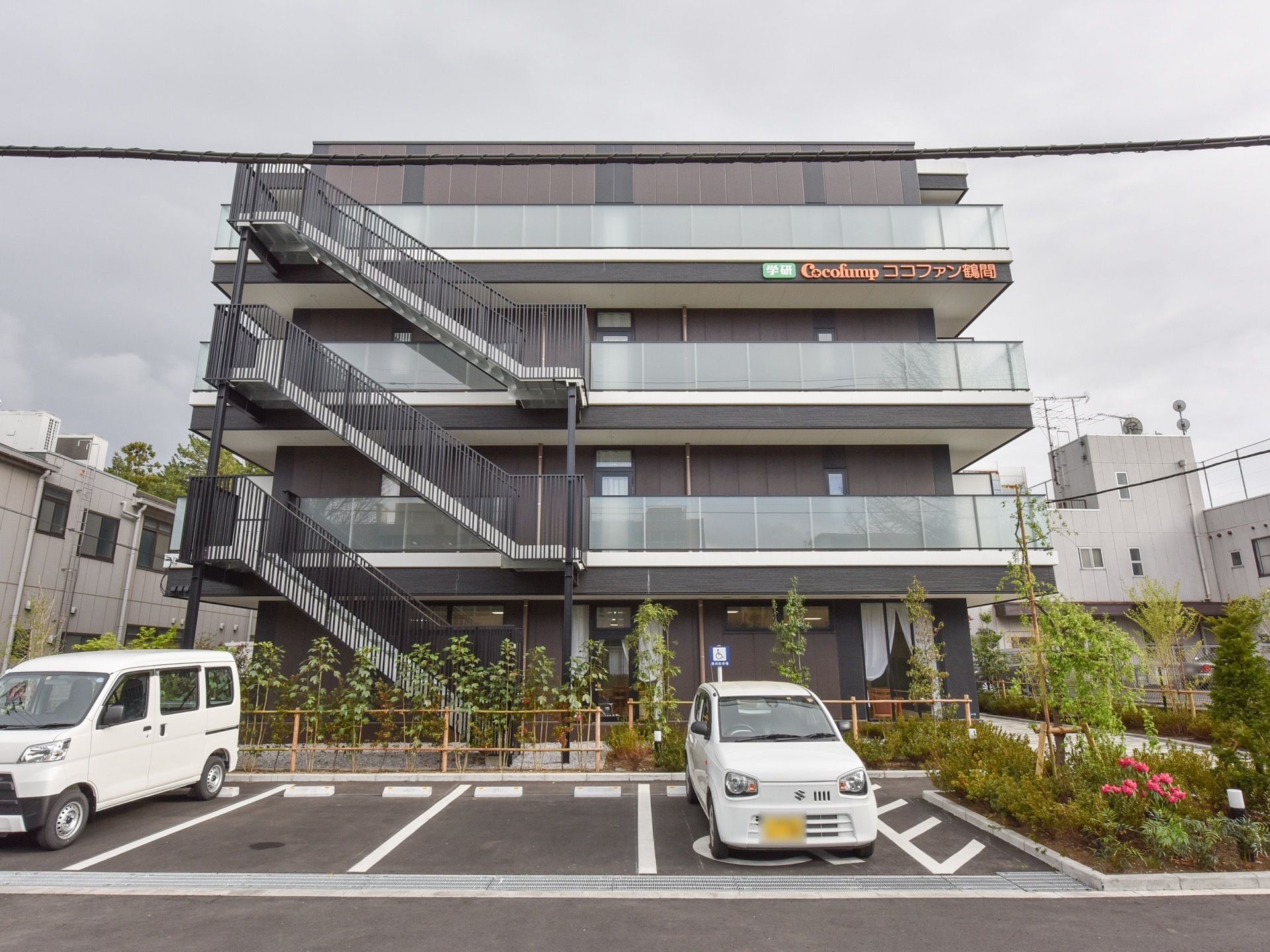 施設の外観。4階まである建物で、左手に外階段が付いている。施設前に植物が植えられているスペースがあり、駐車場がある。