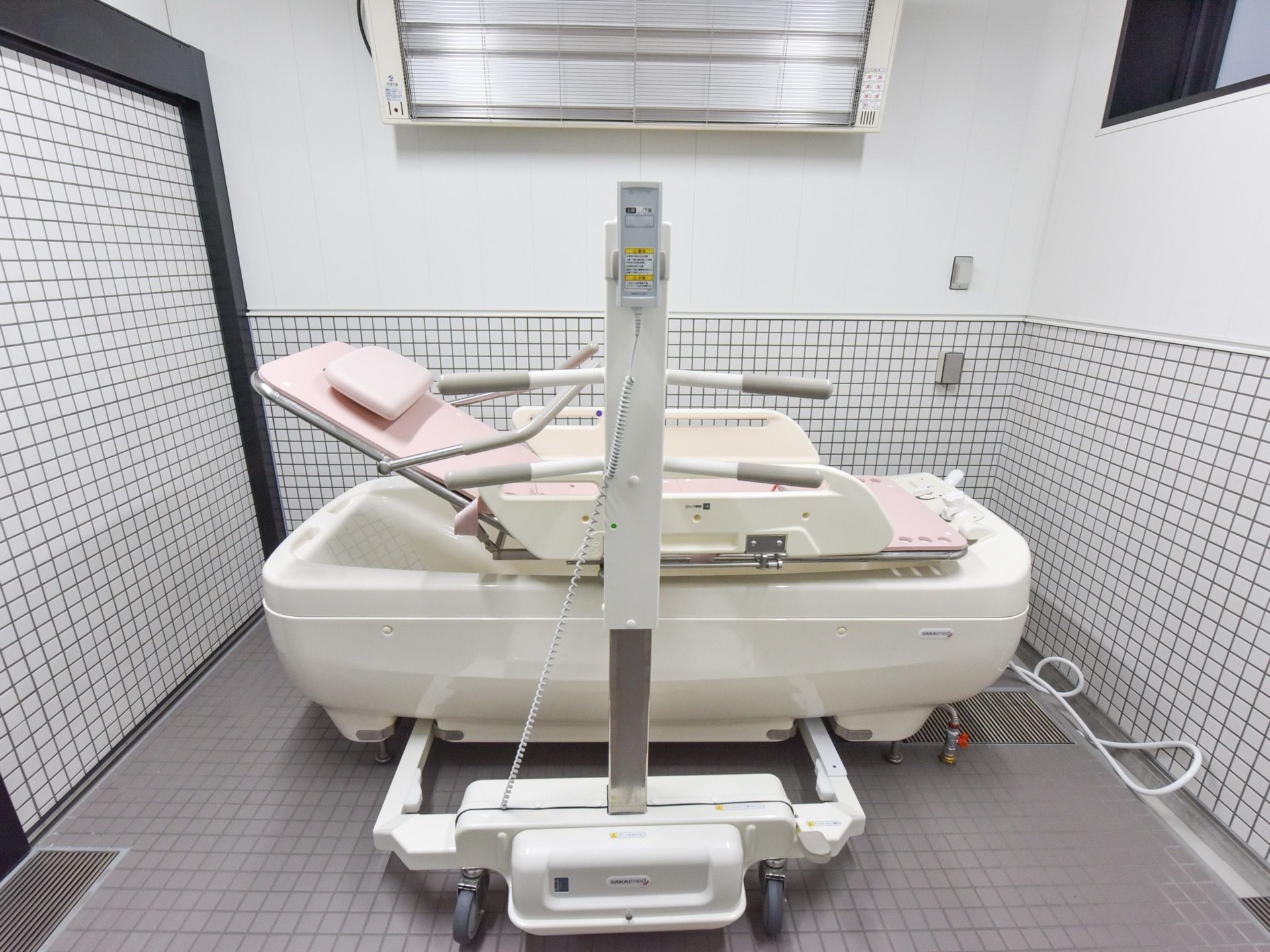 浴室内に、入浴機器が置かれている。横長の浴槽があり、キャスター付きの機器がその前にある。横になったまま入浴できるタイプの機器である。