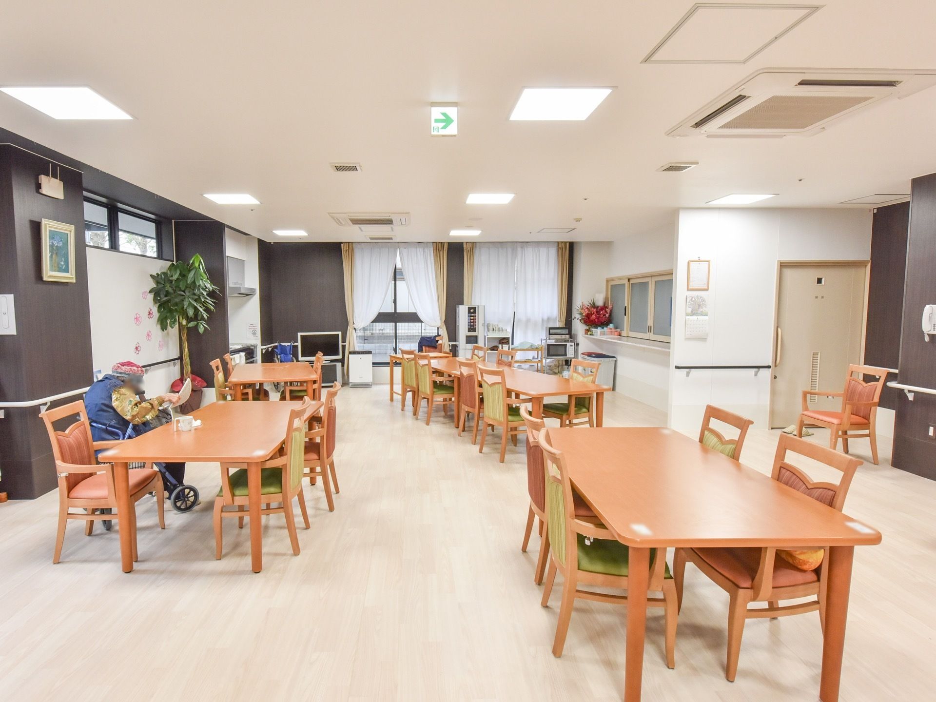 テーブルセットが5つあり、1人の入居者様が車椅子に座っている。左奥にテレビがあり、右奥には給水器や電子レンジがある。
