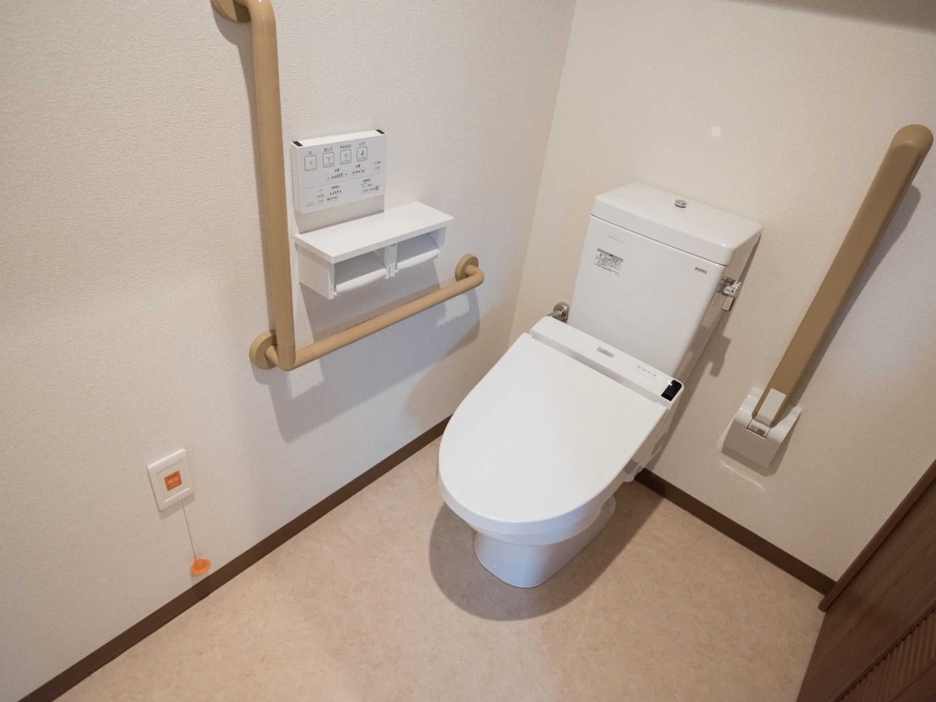 清潔感があり、モダンな印象のトイレである。温水洗浄便座が設置され、Ｌ字型の手すりと可動式の手すり、緊急ボタンが備え付けられている。