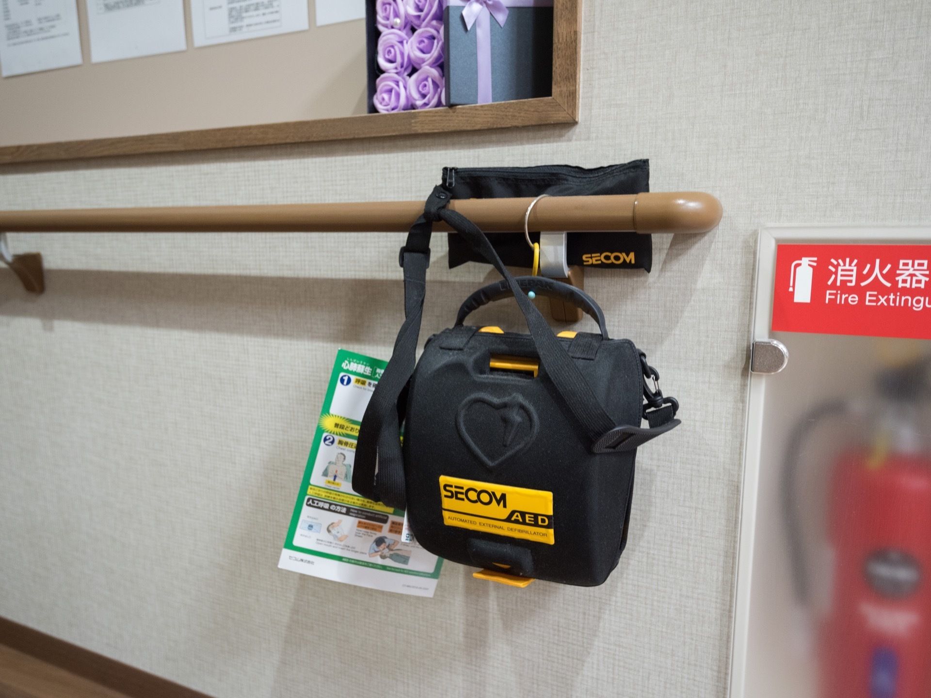 黒いカバーに入ったAEDが、手すりに吊り下げられている。また、壁に収納されるかたちで消火器が設置されている。