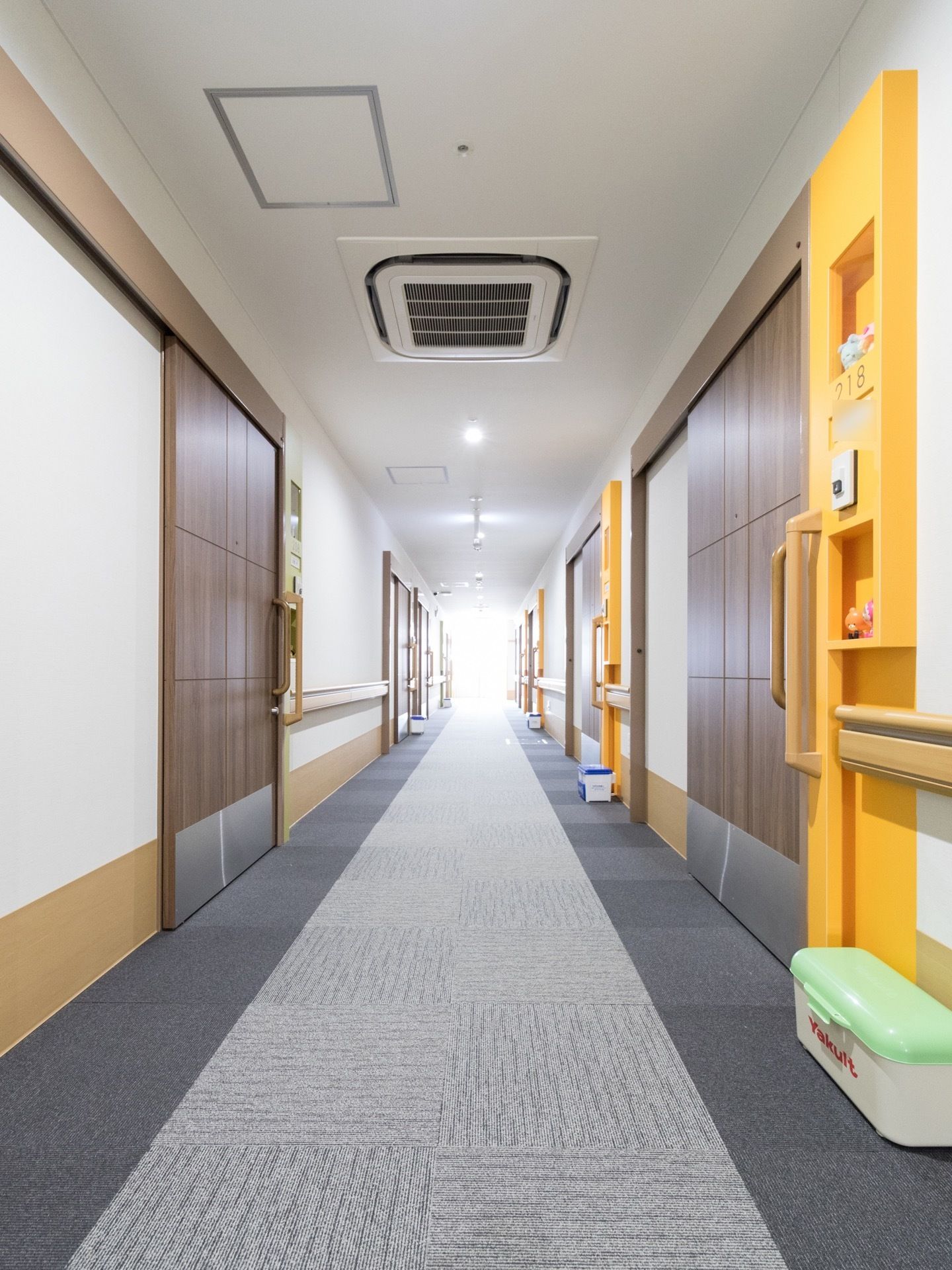 幅が広い廊下。両側の壁には手すりがつけられており、居室の前には牛乳やヤクルトの宅配用の箱がおかれている。